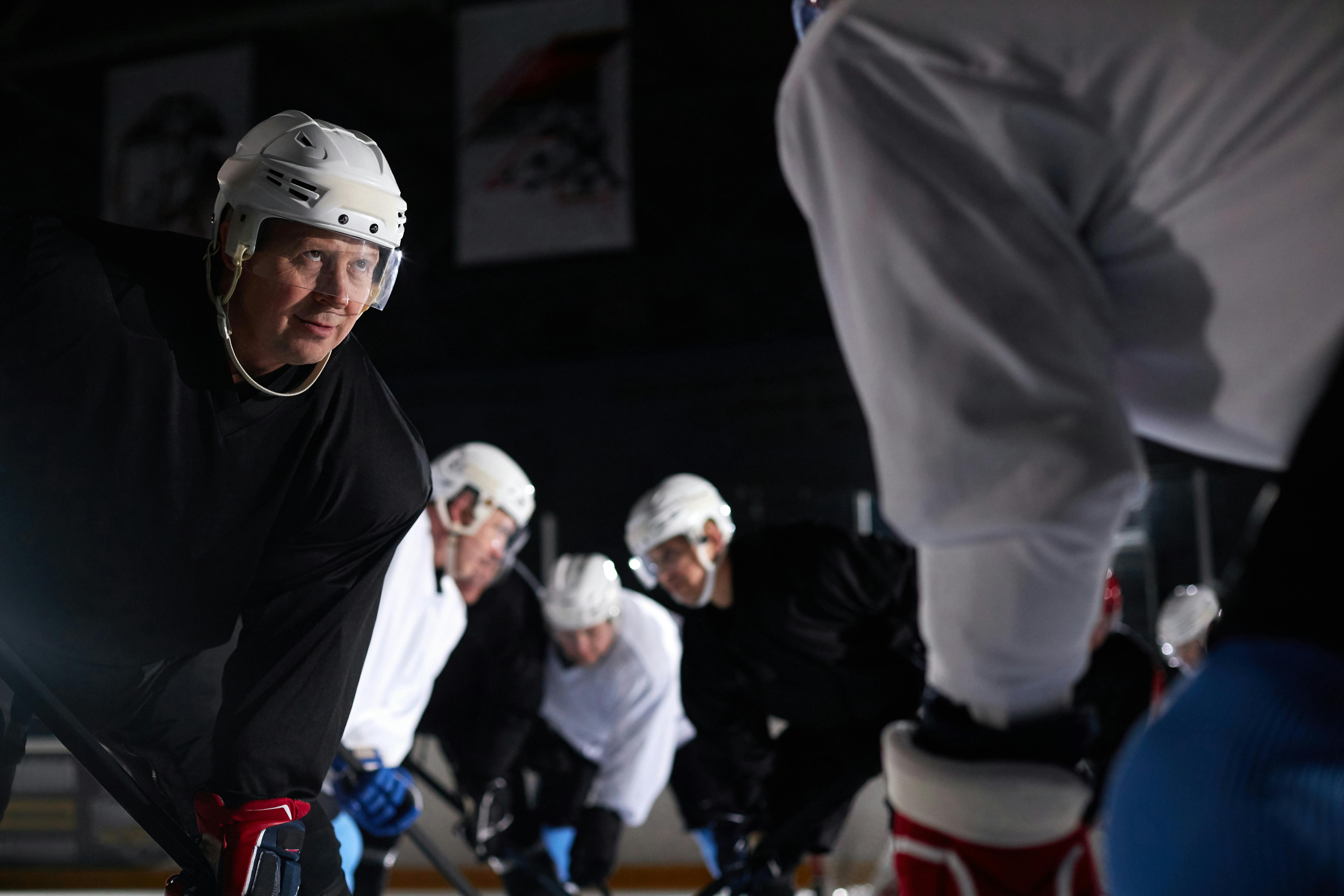 Adult male hockey players in full gear during a faceoff on an indoor ice rink.